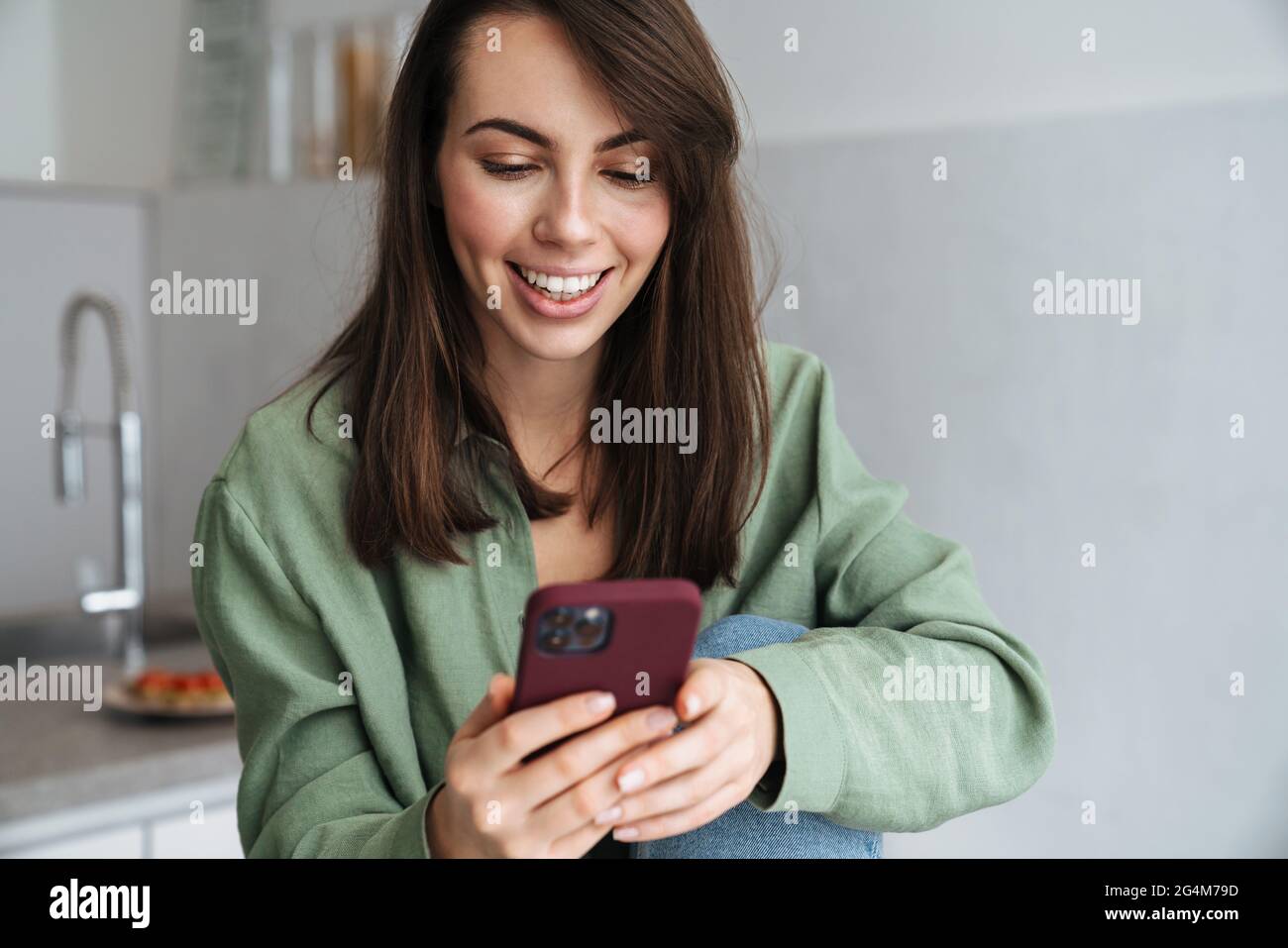 Young smiling woman using mobile phone while sitting at home kitchen Stock Photo