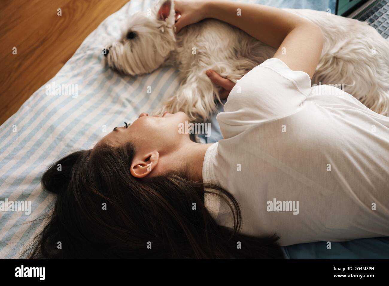 Happy white woman stroking her dog while lying on bed at home Stock