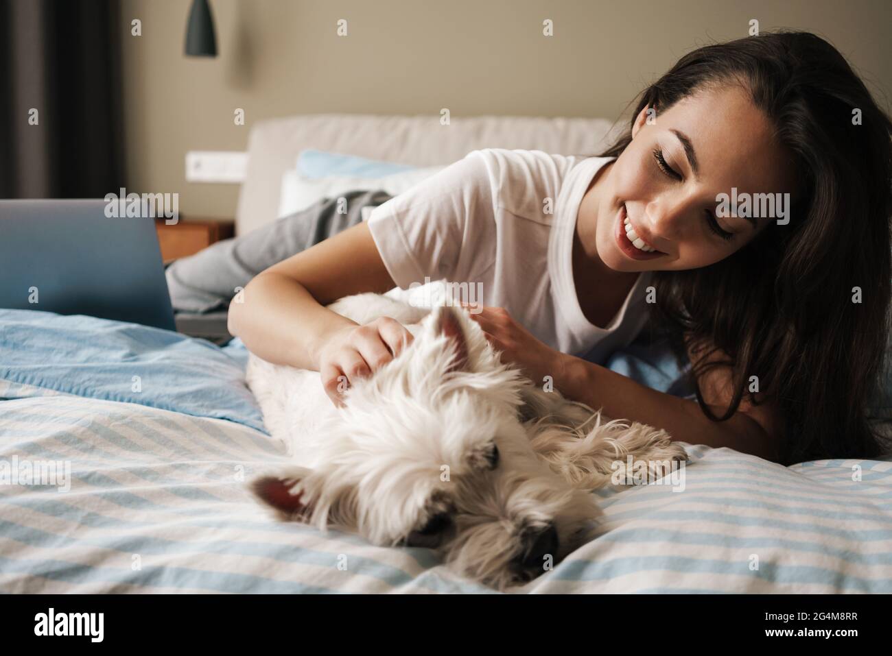 Happy white woman stroking her dog while lying on bed at home Stock