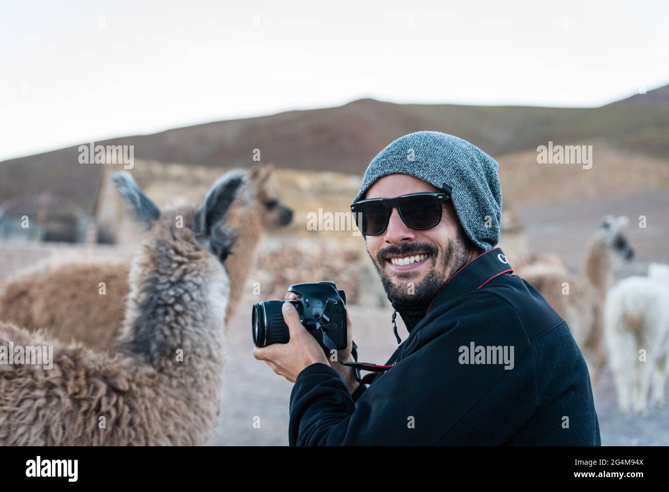 A young man wearing sunglasses and a hut smiles next to a llama with his camera in the Andean altiplano, in Bolivia. Stock Photo