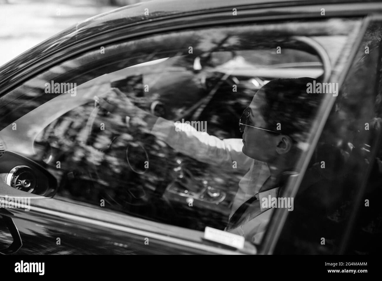 Beautiful young woman sits behind the wheel in black modern car. View through glass in side window. Black and white image. Stock Photo