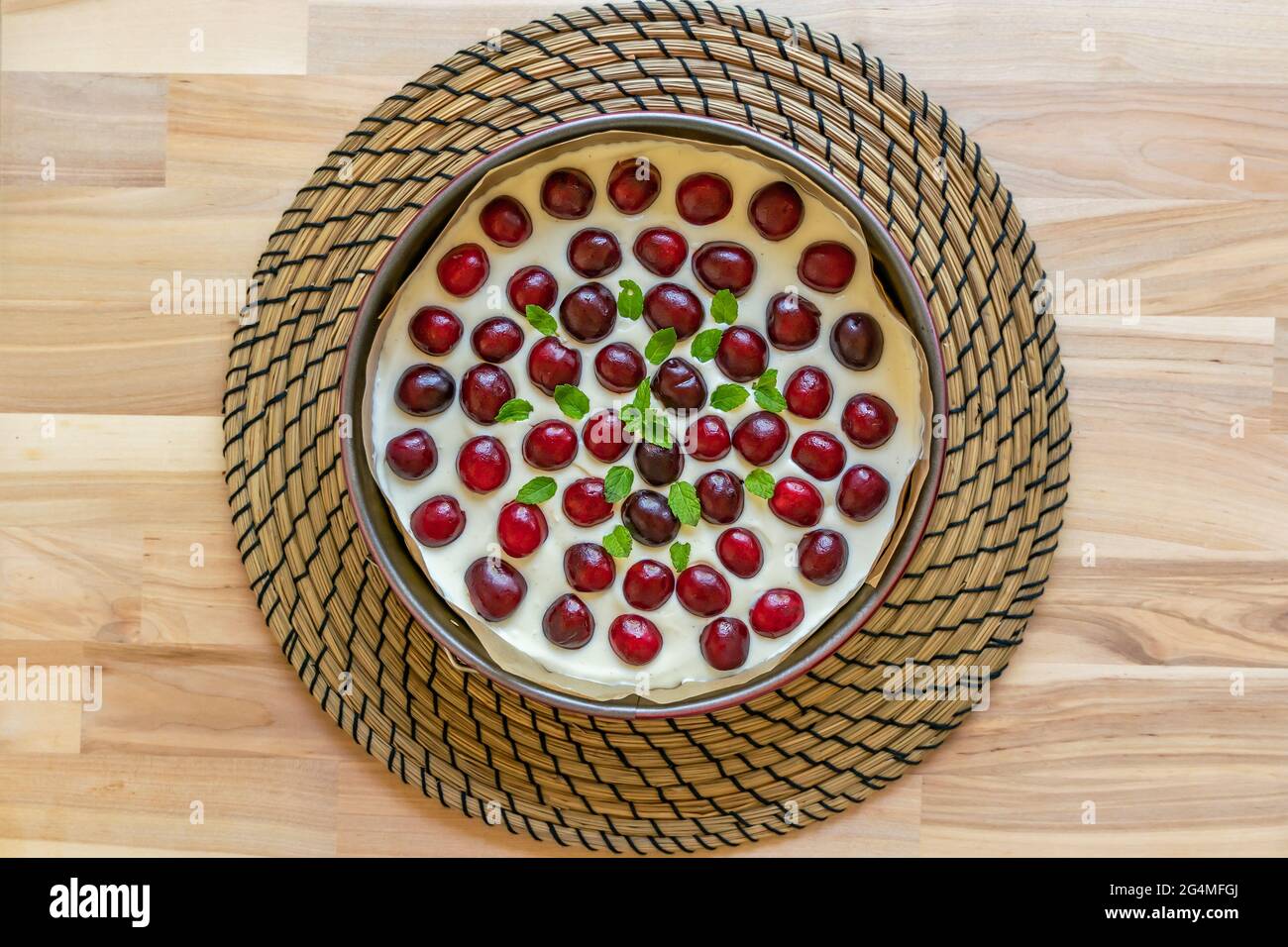 Homemade cheesecake is topped with fresh cherries and a sprig of mint in round cake pan on wooden table Stock Photo