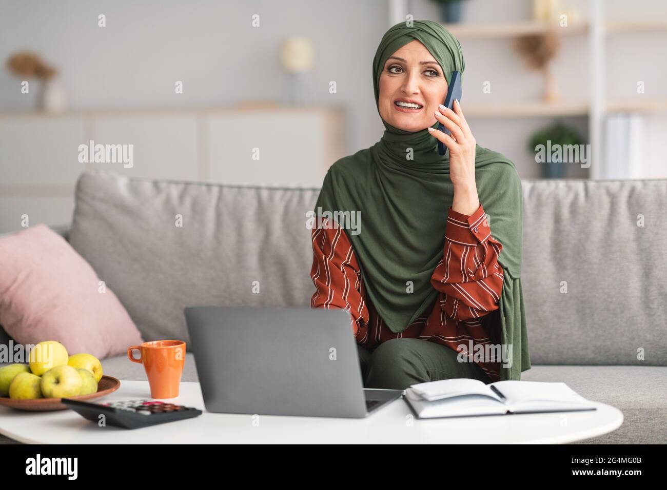 Mature Muslim Woman Talking On Phone Using Laptop At Home Stock Photo