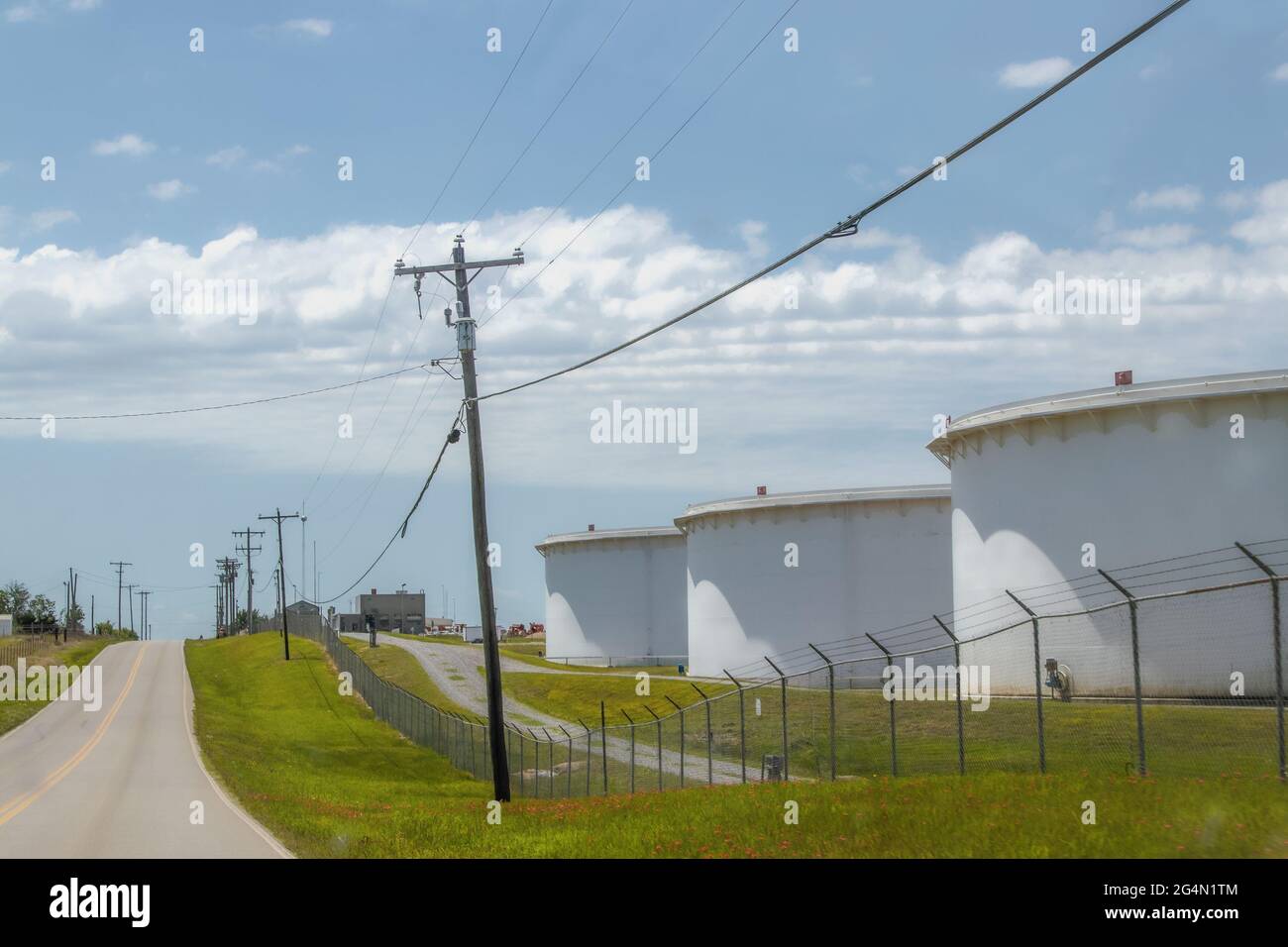 Huge oil containers at tank farm behind chain link fence by country blacktop road with powerlines in Cushing OK USA Stock Photo