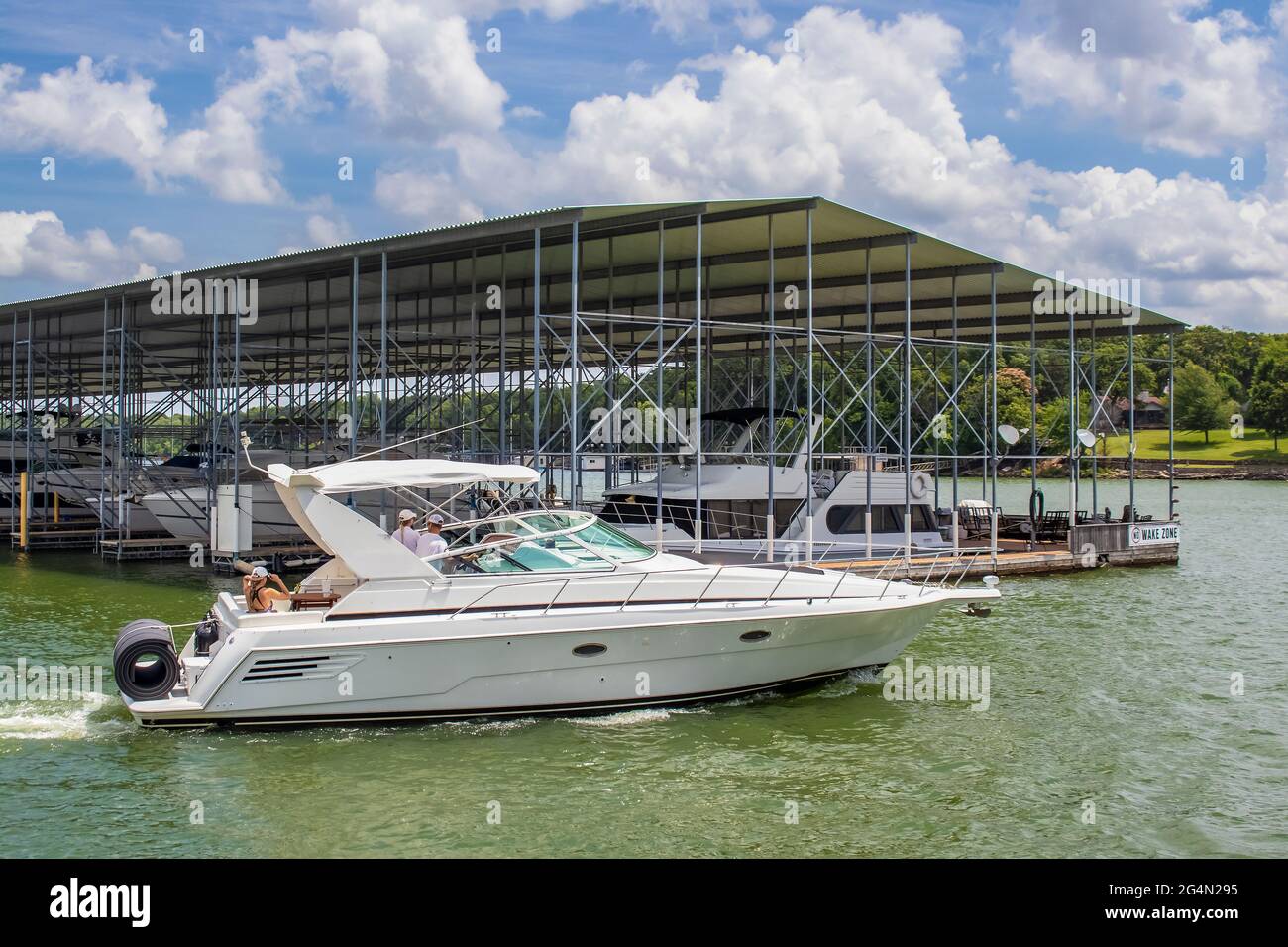 Luxury speedboat with rolled floatie on back on lake in summer passing by covered boat dock with more fancy boats and No Wake Zone sign Stock Photo