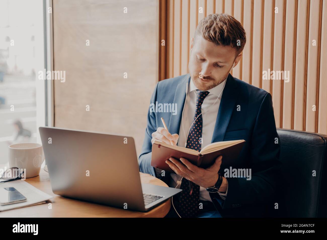 Concentrated entrepreneur in formal outfit wearing earphones watching online webinar on laptop in cafe Stock Photo