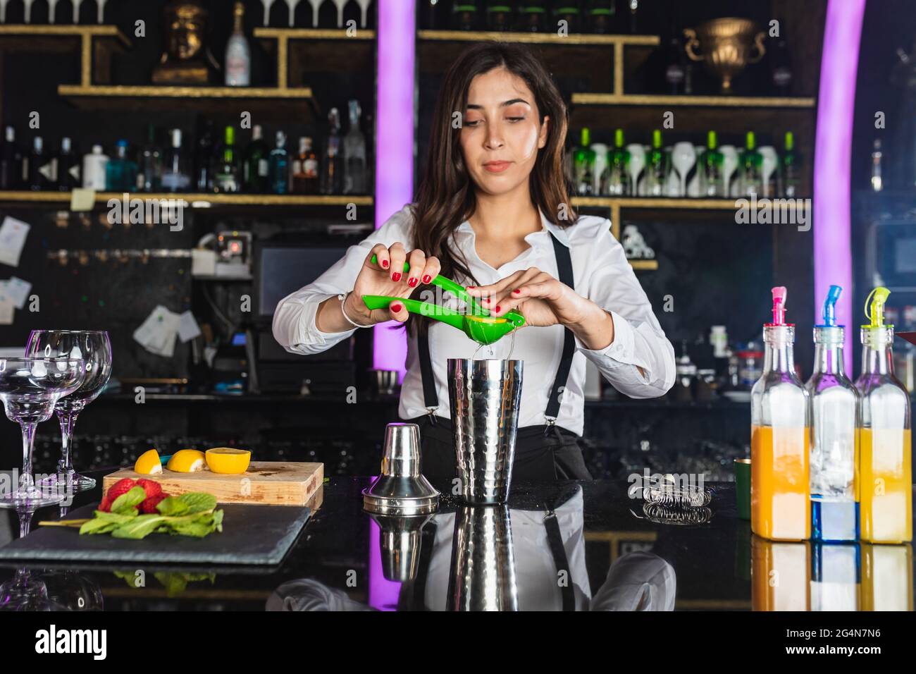 Young female barkeeper in stylish outfit squeezing lemon while preparing cocktail standing at counter in modern bar Stock Photo