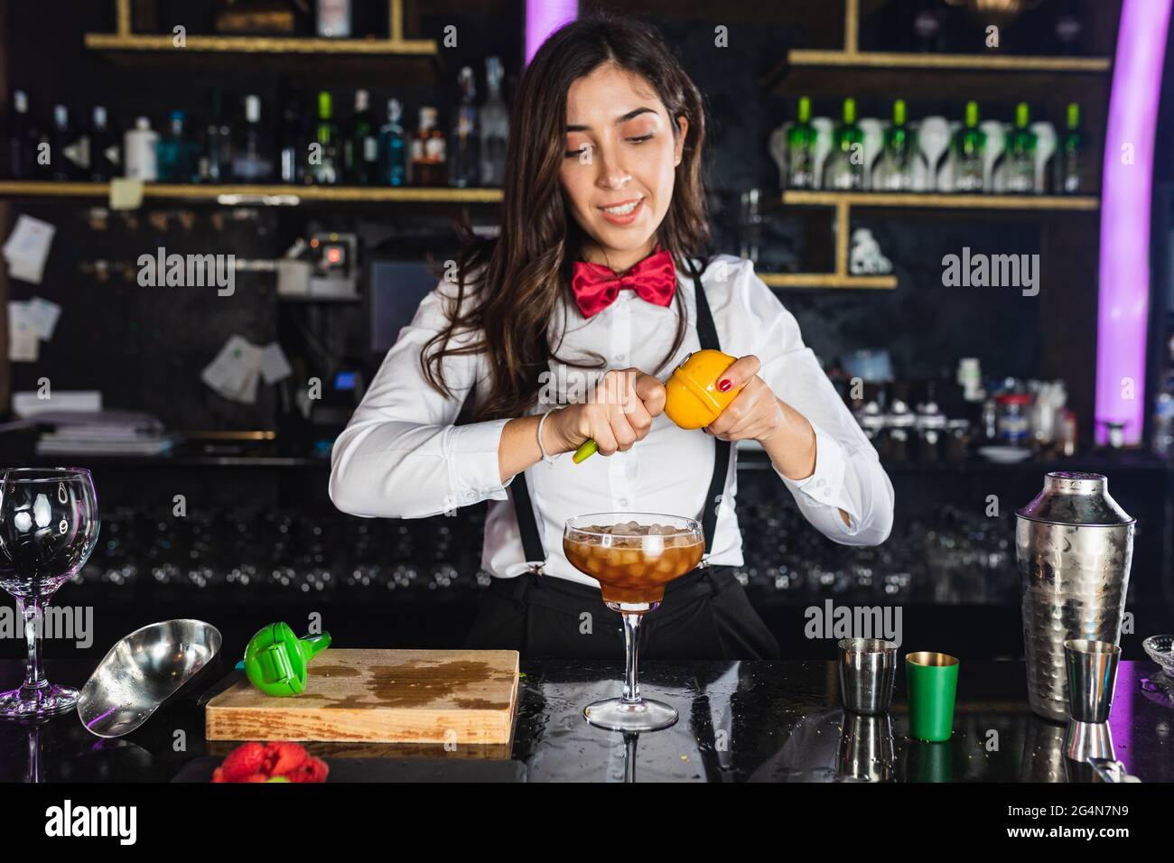 Young female barkeeper in stylish outfit decorating cocktail with lemon peel while standing at counter in modern bar Stock Photo
