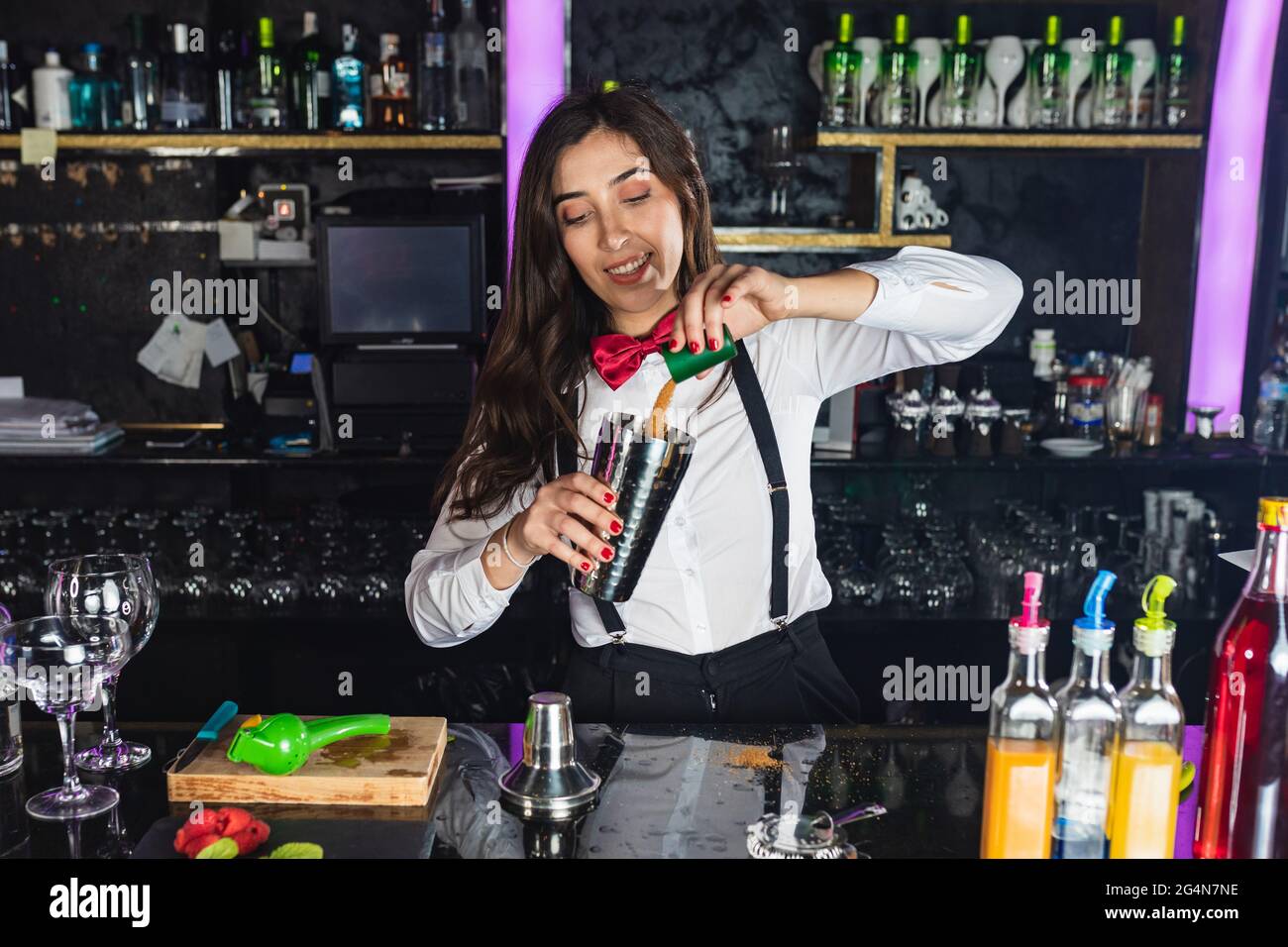 Happy female barkeeper in stylish outfit adding sugar into shaker while preparing cocktail standing at counter in modern bar Stock Photo