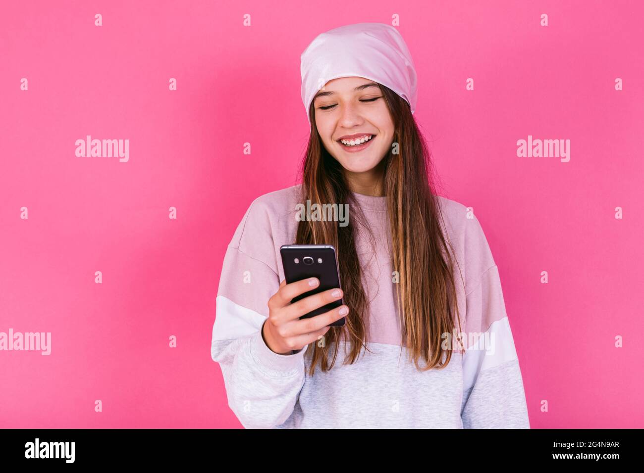 Long haired blond Hispanic female model wearing dark leather jacket and cap while smiling and looking at camera on pink backdrop in studio Stock Photo