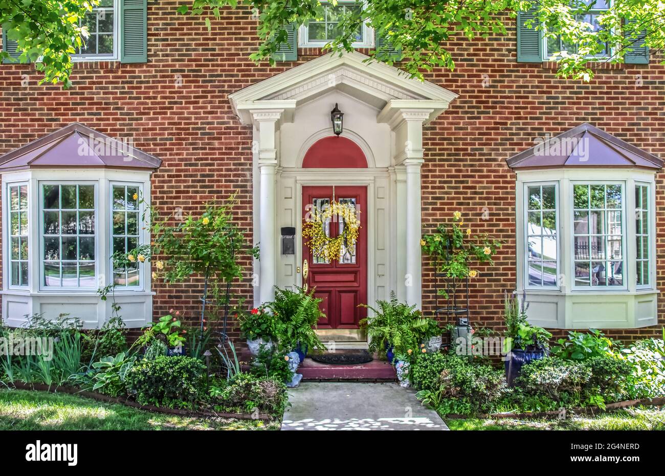Entrance with columns and bay windows of beautiful two-story brick house with pots of flowers and yellow climbing roses and headplanters and ferns fra Stock Photo