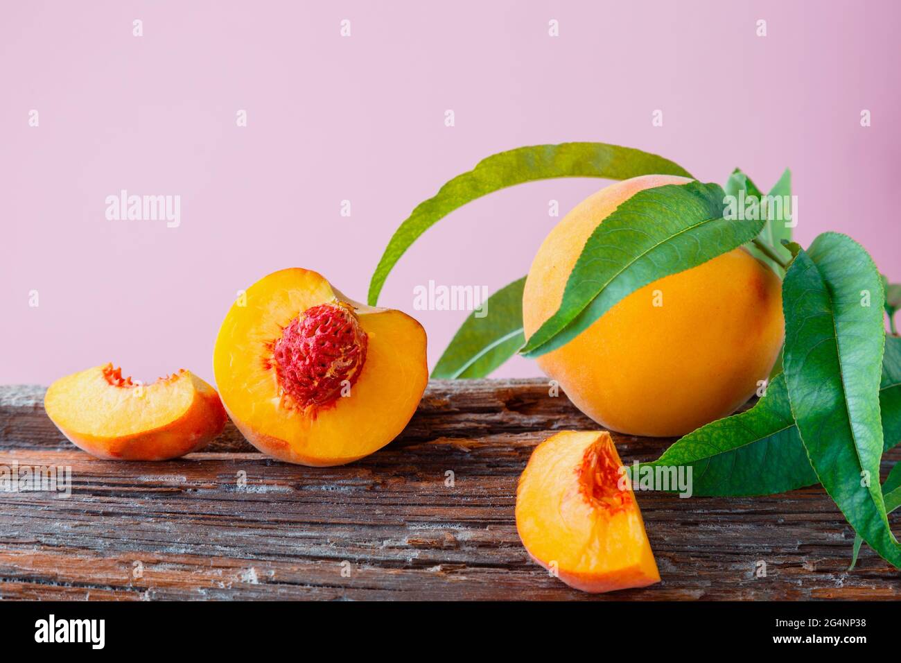 Peaches with leaves on wooden board. Peach in halves with bone, chopped pieces wedges peach halves. Fresh organic peaches fruit on color pink Stock Photo