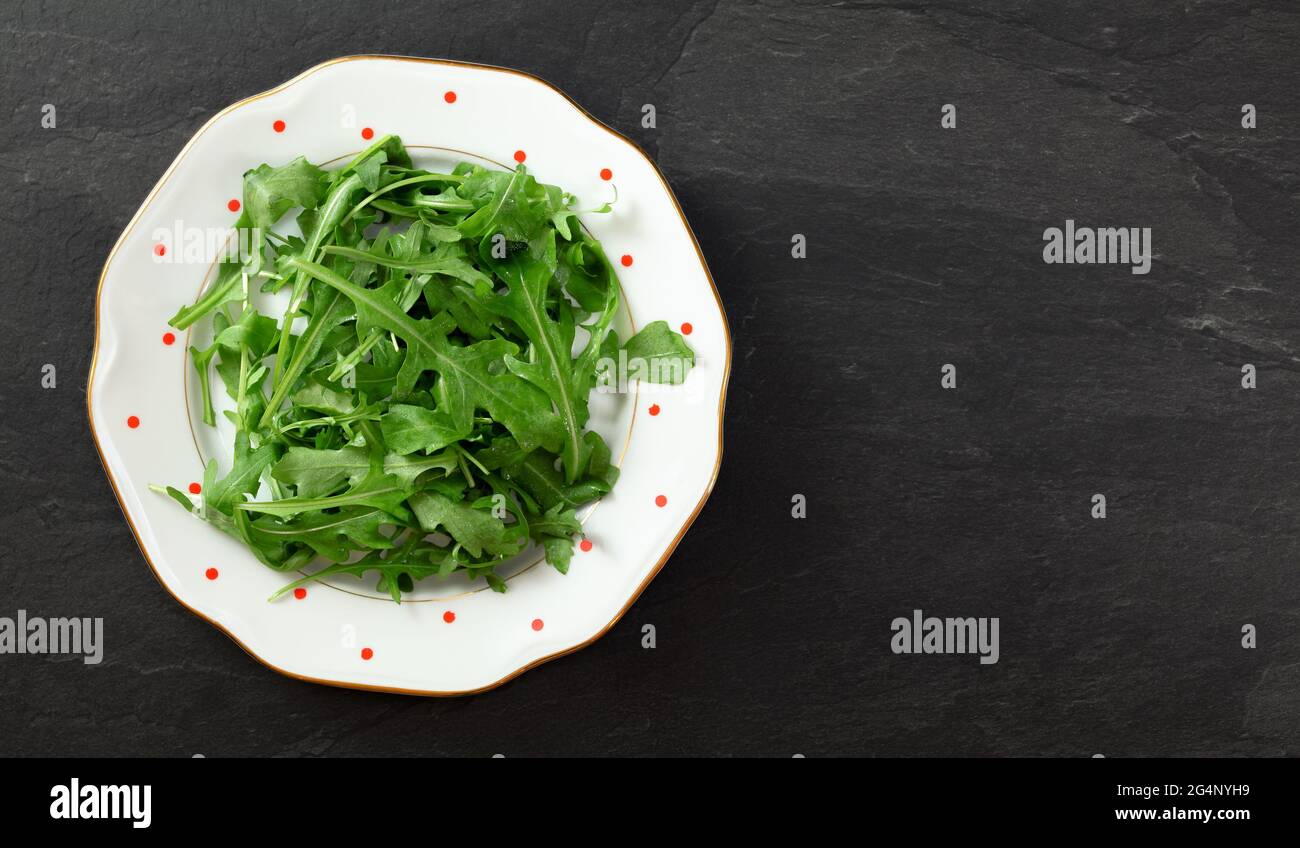 Fresh green arugula or garden rocket leaves on white plate with red dots, black table (space for text right side) under. Healthy greens and salads con Stock Photo