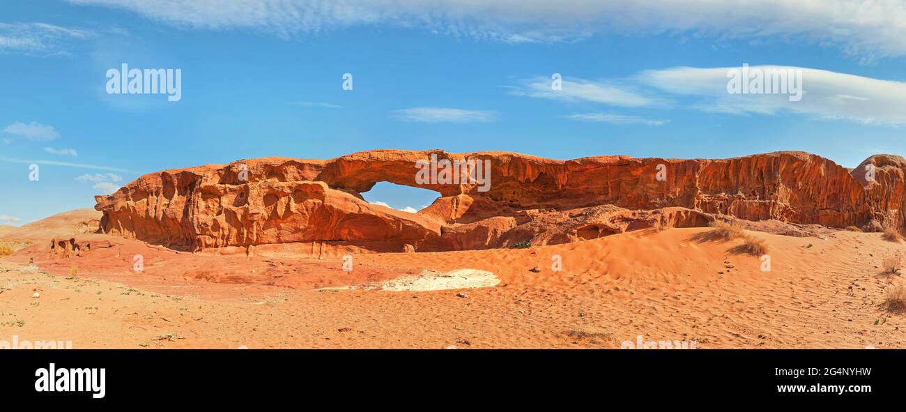 Little arc or small rock window formation in Wadi Rum desert, bright sun shines on red dust and rocks, blue sky above, high resolution wide panorama Stock Photo