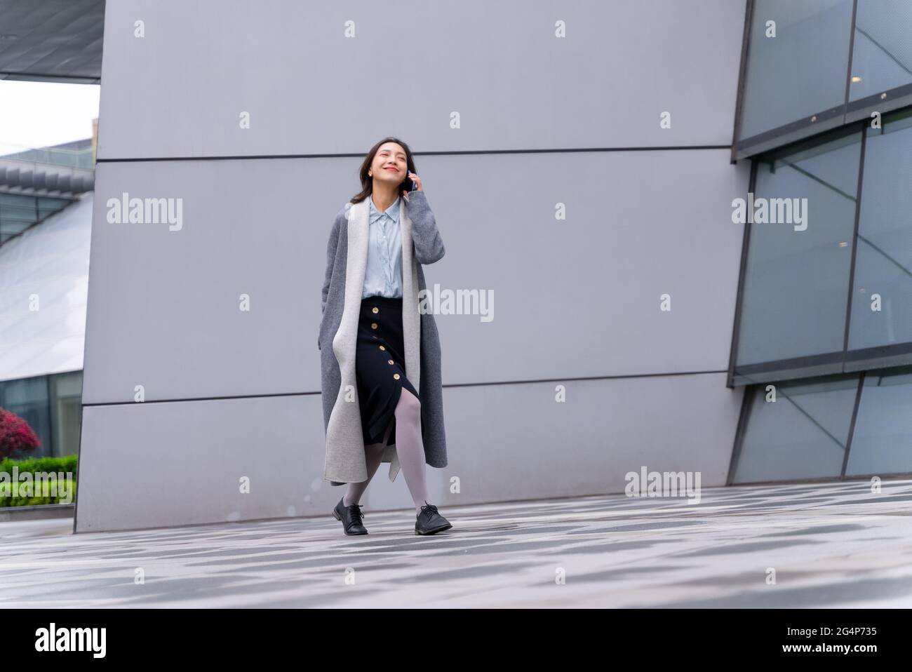 full length view of Young stylish Chinese businesswoman holding mobile phone calling and chatting walking outside modern office building in the Chengd Stock Photo