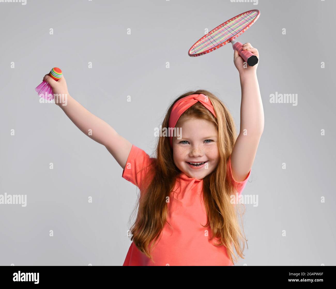 Happy active red-haired kid girl in pink t-shirt and headband holds a badminton racquet and shuttlecock in hands up Stock Photo