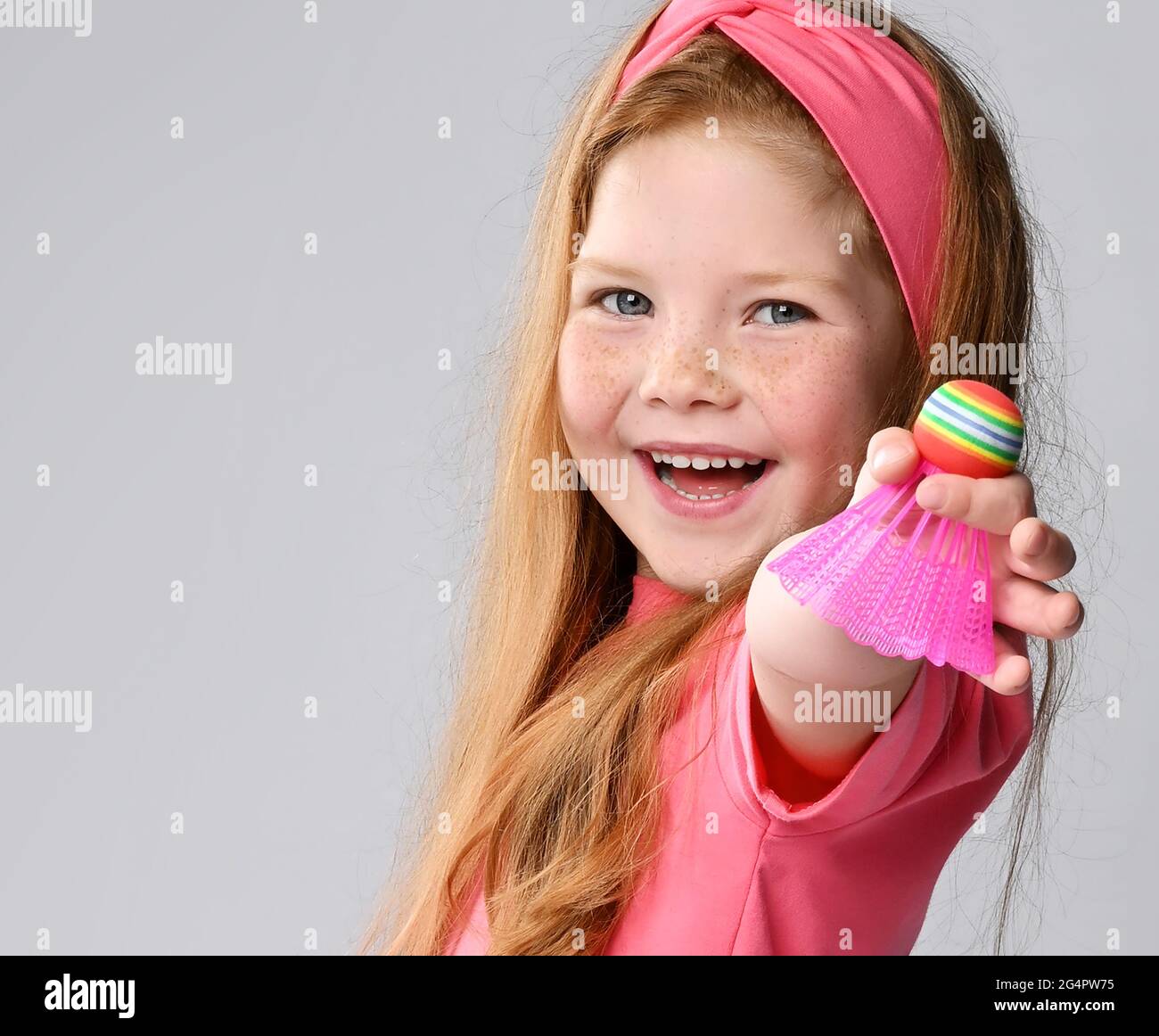 Happy laughing red-haired kid girl in pink t-shirt and headband holds, shows a badminton shuttlecock in hand Stock Photo