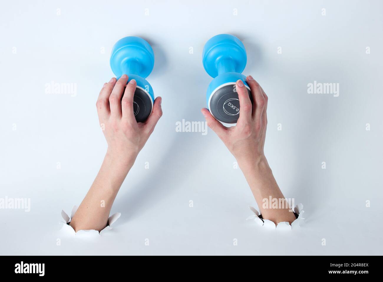 Female hands holding a dumbbell through the torn holes of white background. Creative sports art Stock Photo
