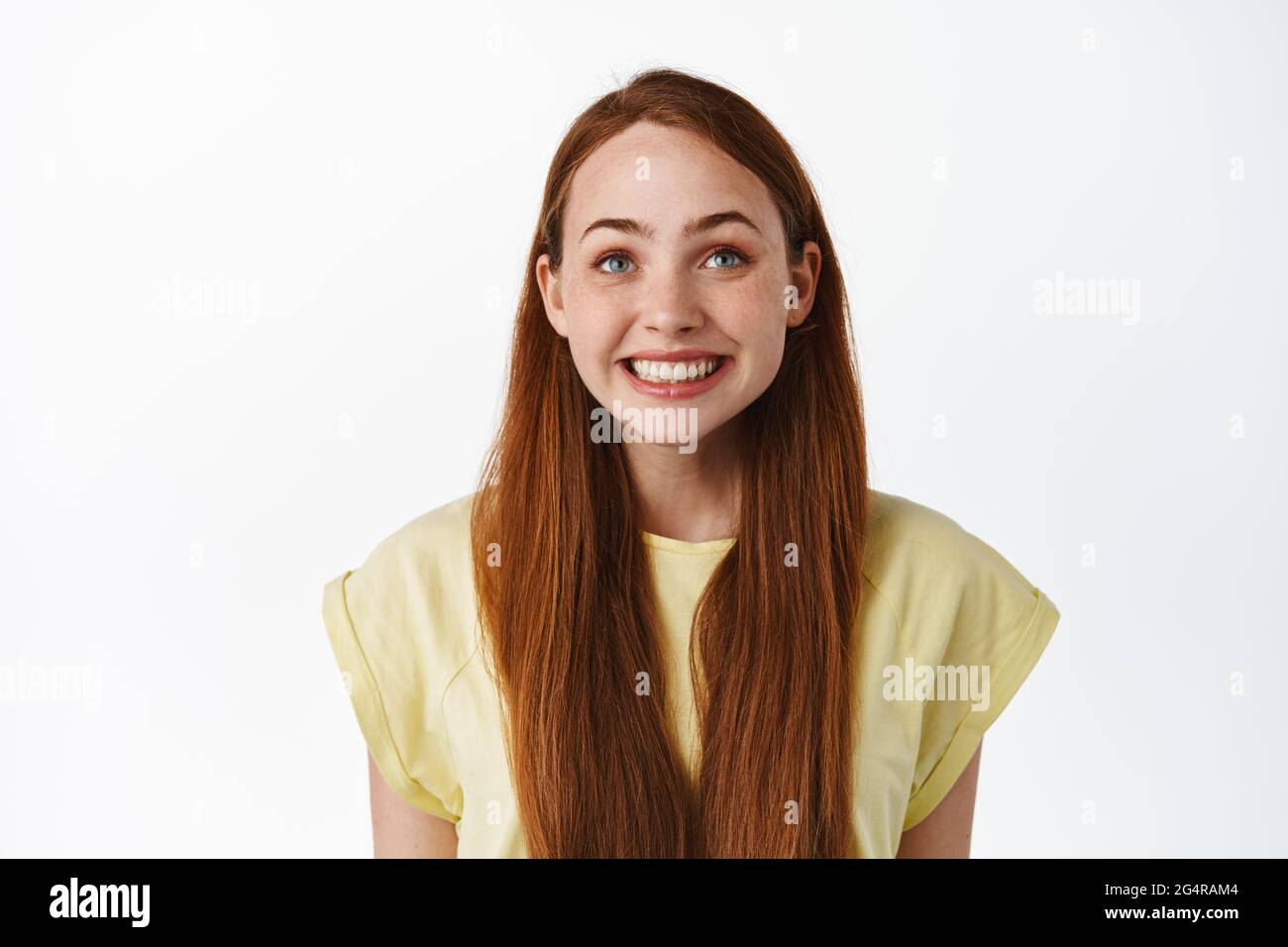 Close up of hopeful pretty girl with long red hair and freckles, looking below camera at tv screen, smiling happy and pleased with results, standing Stock Photo
