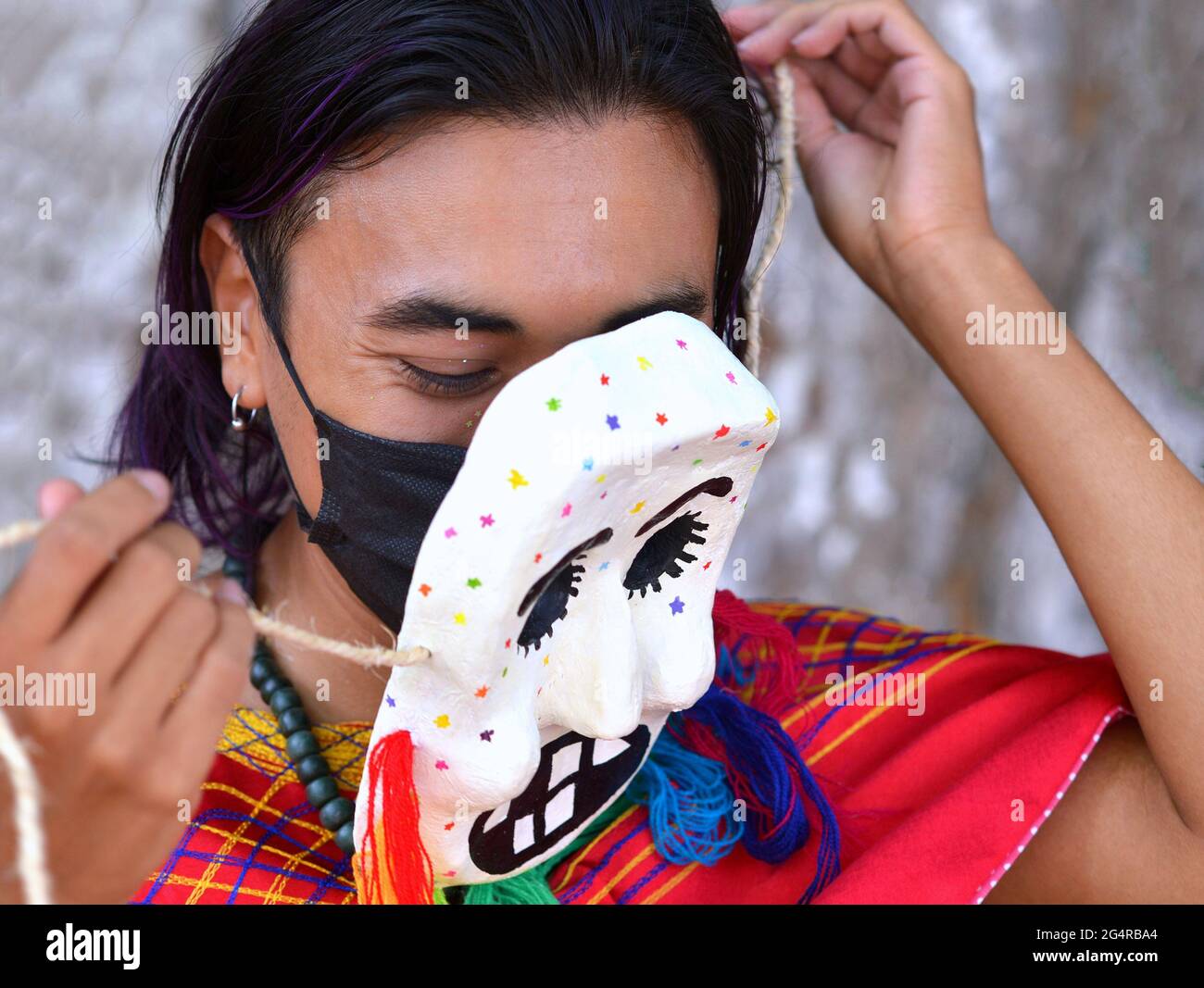 Young Mexican artist (mask maker, mask painter) removes one of his hand painted papier-mache masks and shows a black surgical mask underneath. Stock Photo