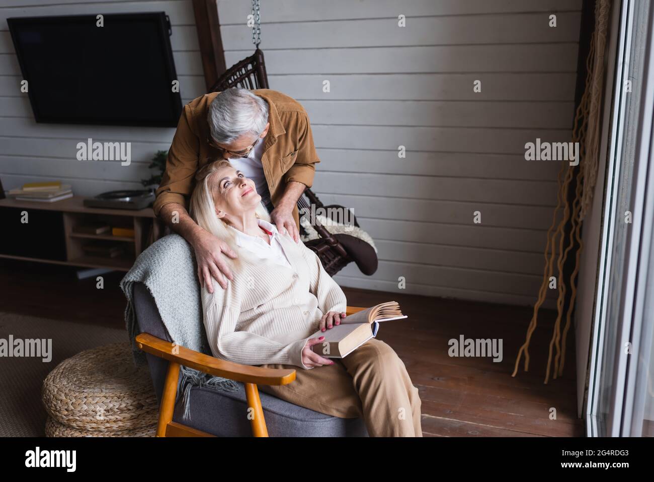 Elderly man hugging smiling wife with book in armchair at home Stock Photo