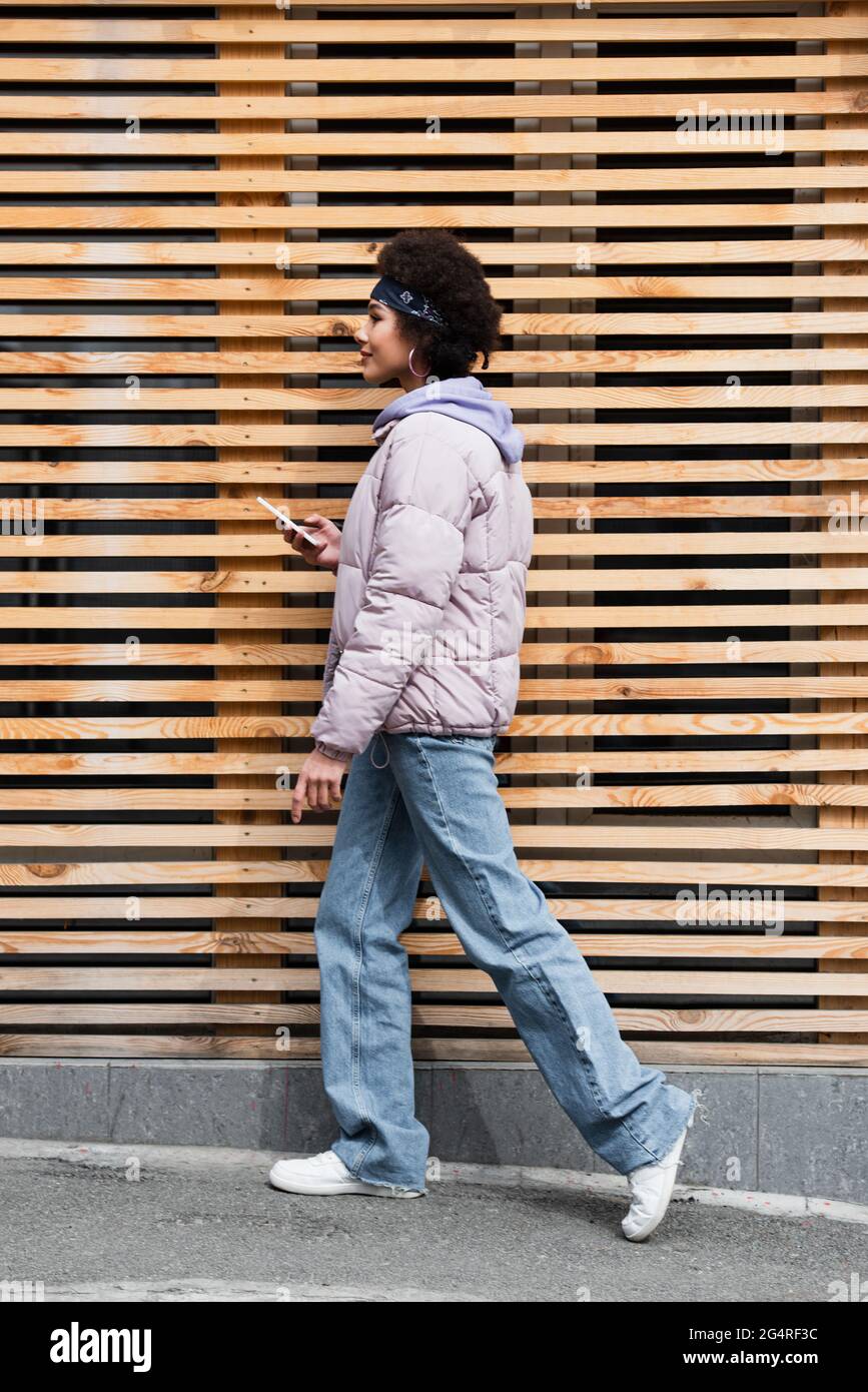 Side view of trendy african american woman with smartphone walking near ...