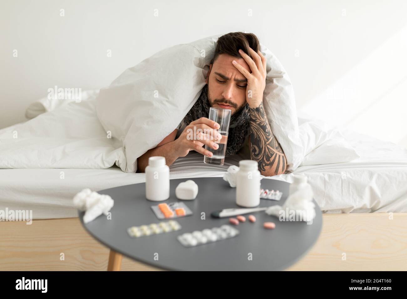 Sick man touching forehead and drinking water, having fever during influenza illness, lying in bed under blanket. Covid-19 coronavirus and seasonal fl Stock Photo