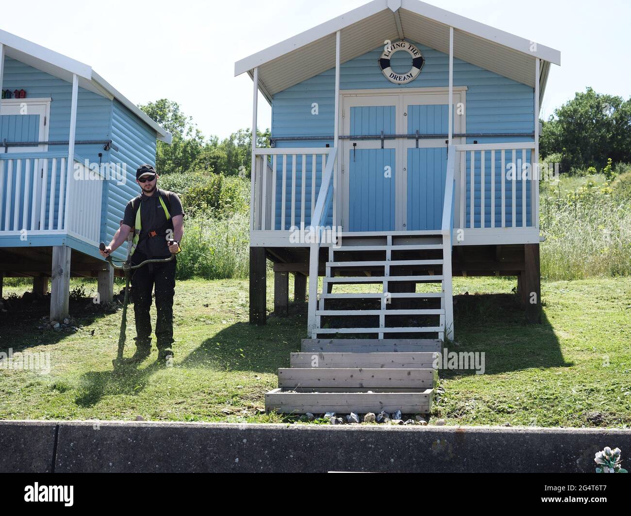 Minster on Sea, Kent, UK. 23rd June, 2021. UK Weather: a sunny & warm afternoon in  Minster on Sea, Kent. A council worker strims the grass in front of a beach hut called 'Living the Dream'. Credit: James Bell/Alamy Live News Stock Photo