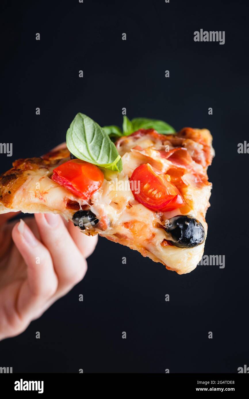Slice of pizza with tomato, melted cheese and olives in female hand isolated on black background Stock Photo