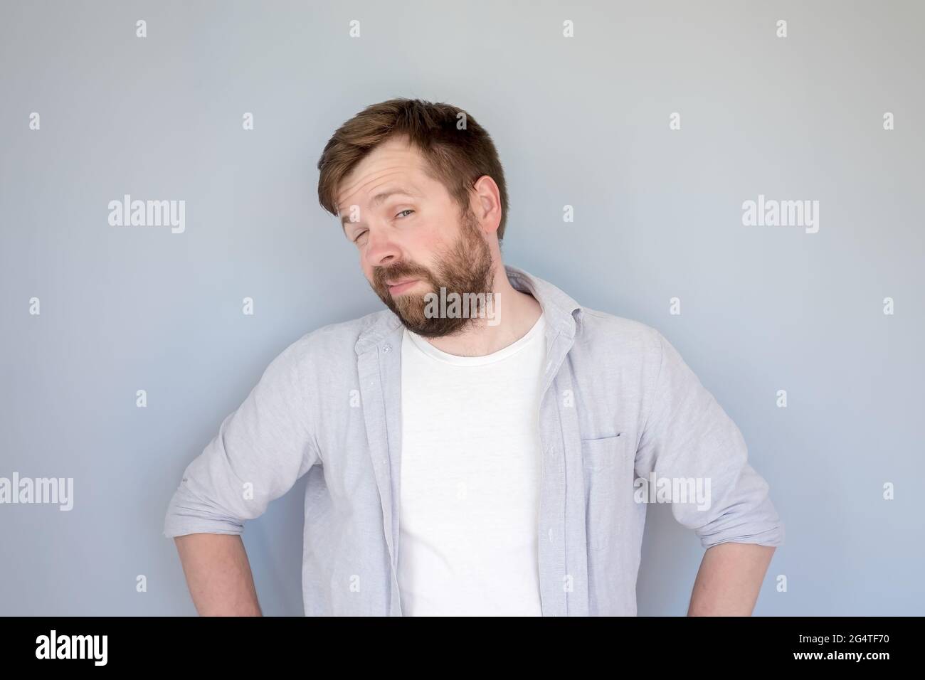 Caucasian bearded man in a shirt looks suspiciously and slyly, narrowing his eyes and akimbo. Gray background. Stock Photo