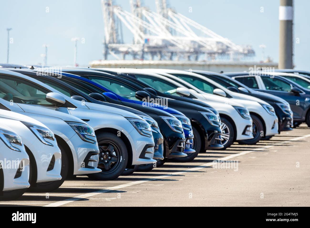 Row of brand new cars lined up outdoors in a parking lot with container cranes in the distance. Stock Photo