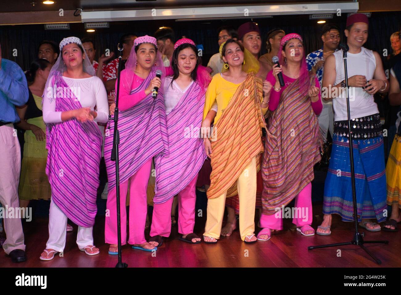 A group of six Filipina ladies in saris singing to an audience on board a cruise ship with Filipino men standing behind Stock Photo