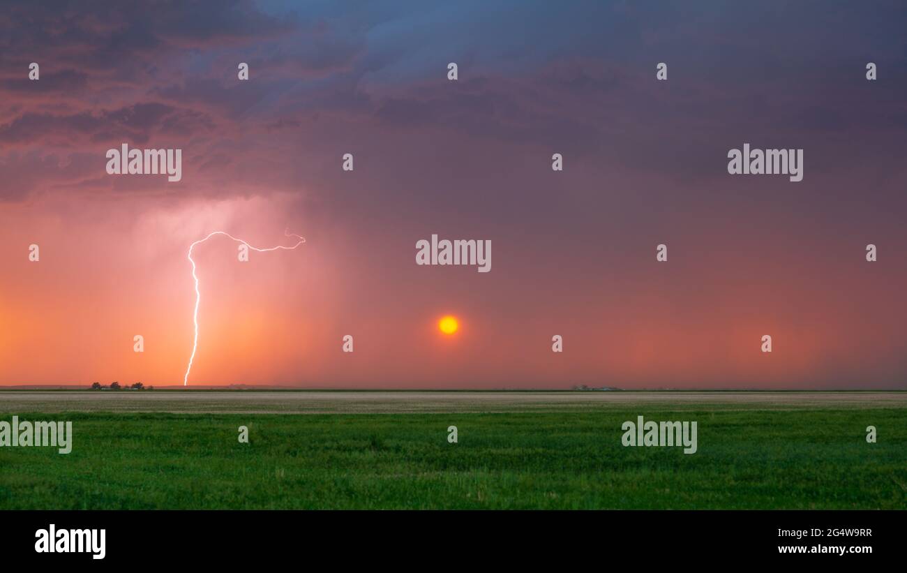 Lightning bolt strikes at sunset with the sun seen heavily filtered by the rain falling in the distance. Dark storm clouds can be seen overhead. Stock Photo