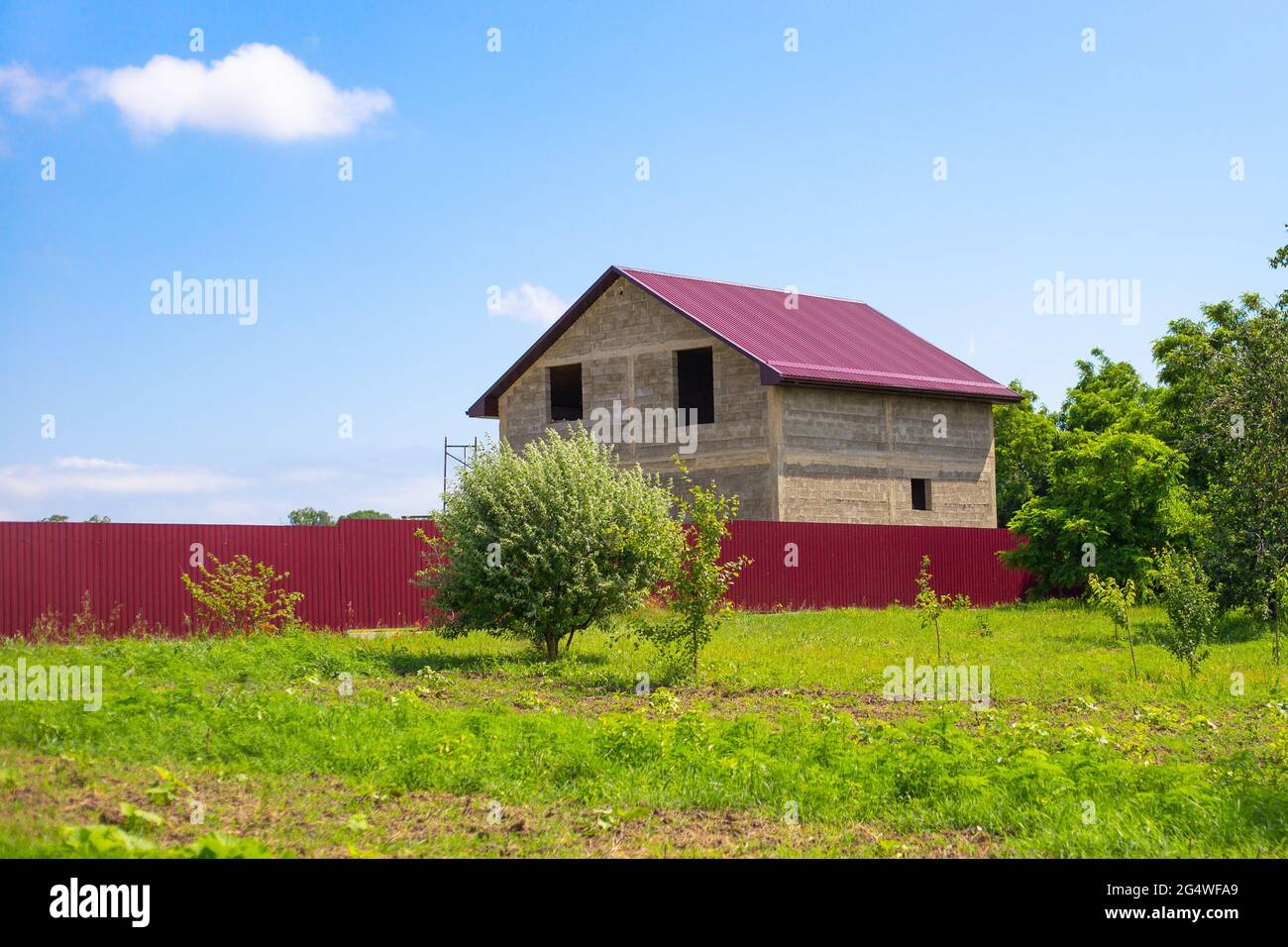 Private house under construction behind a fence. In the foreground is a green garden. Countryside and construction. Stock Photo