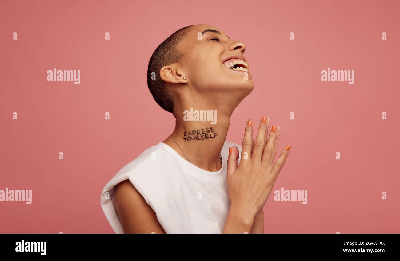 female with shaved head smiling on pink background. Androgynous woman with express yourself written on neck. Stock Photo