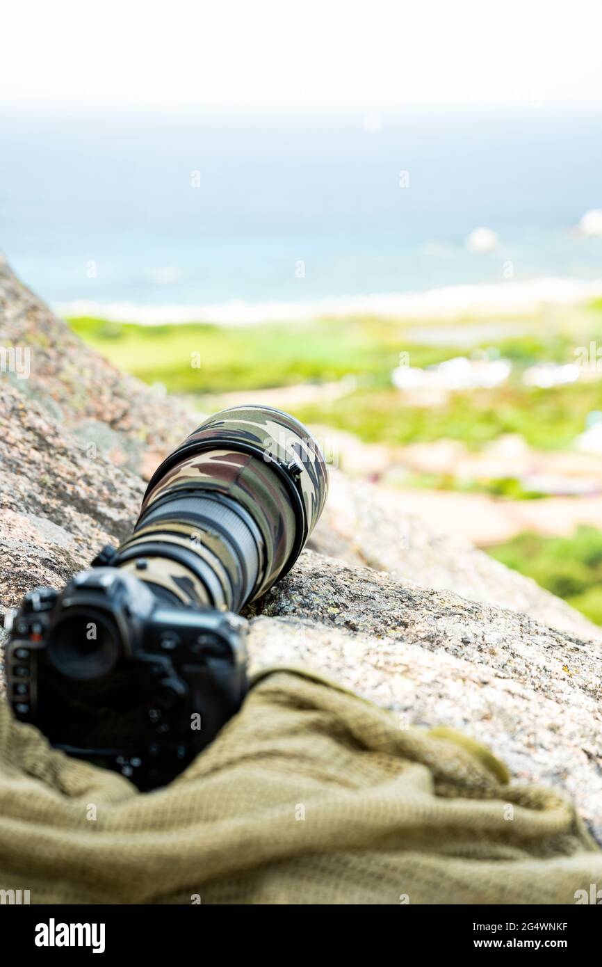 (Selective focus) Professional camera with a super telephoto lens with camouflage cover on a rocky mountains. Paparazzi equipment. Stock Photo