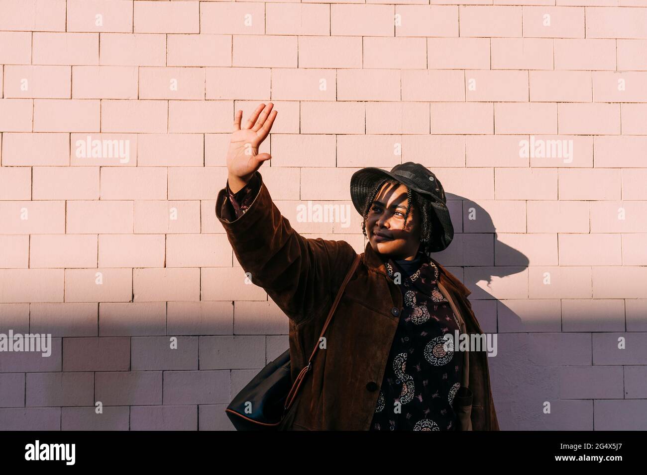 Young woman wearing hat and overcoat stretching hand in front of brick wall Stock Photo