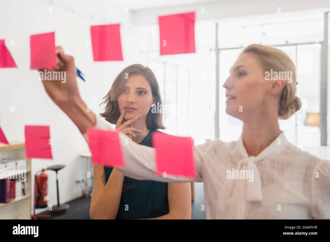 Young female entrepreneur looking at coworker writing on adhesive notes while discussing in office Stock Photo