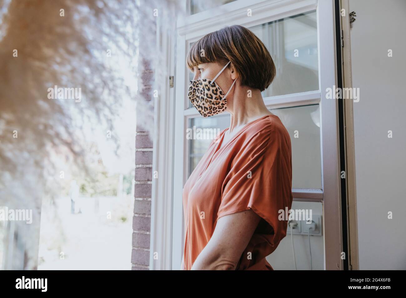 Woman with animal print face mask looking through window at home Stock Photo