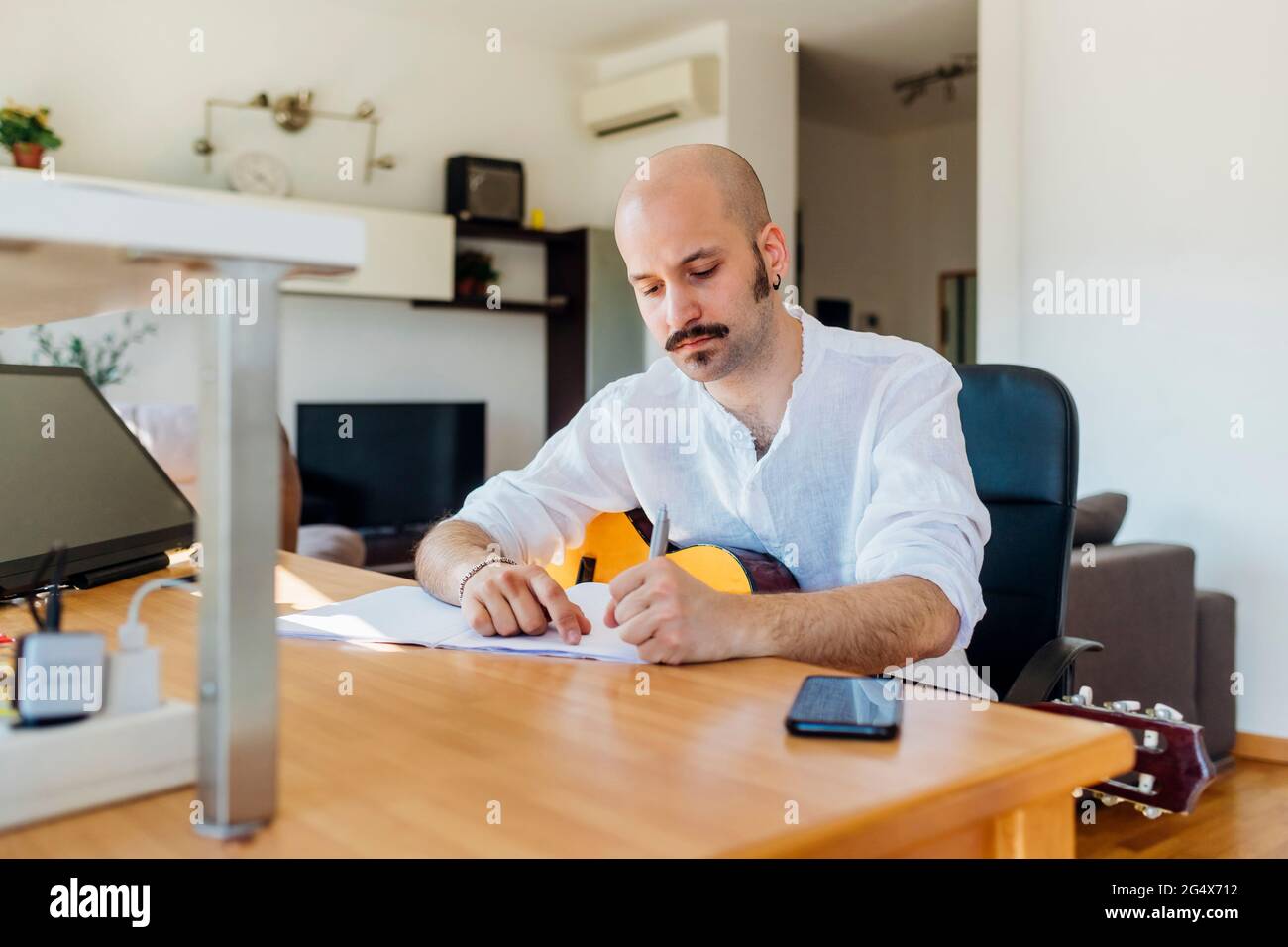 Male music composer with guitar writing musical notes at table in living room Stock Photo