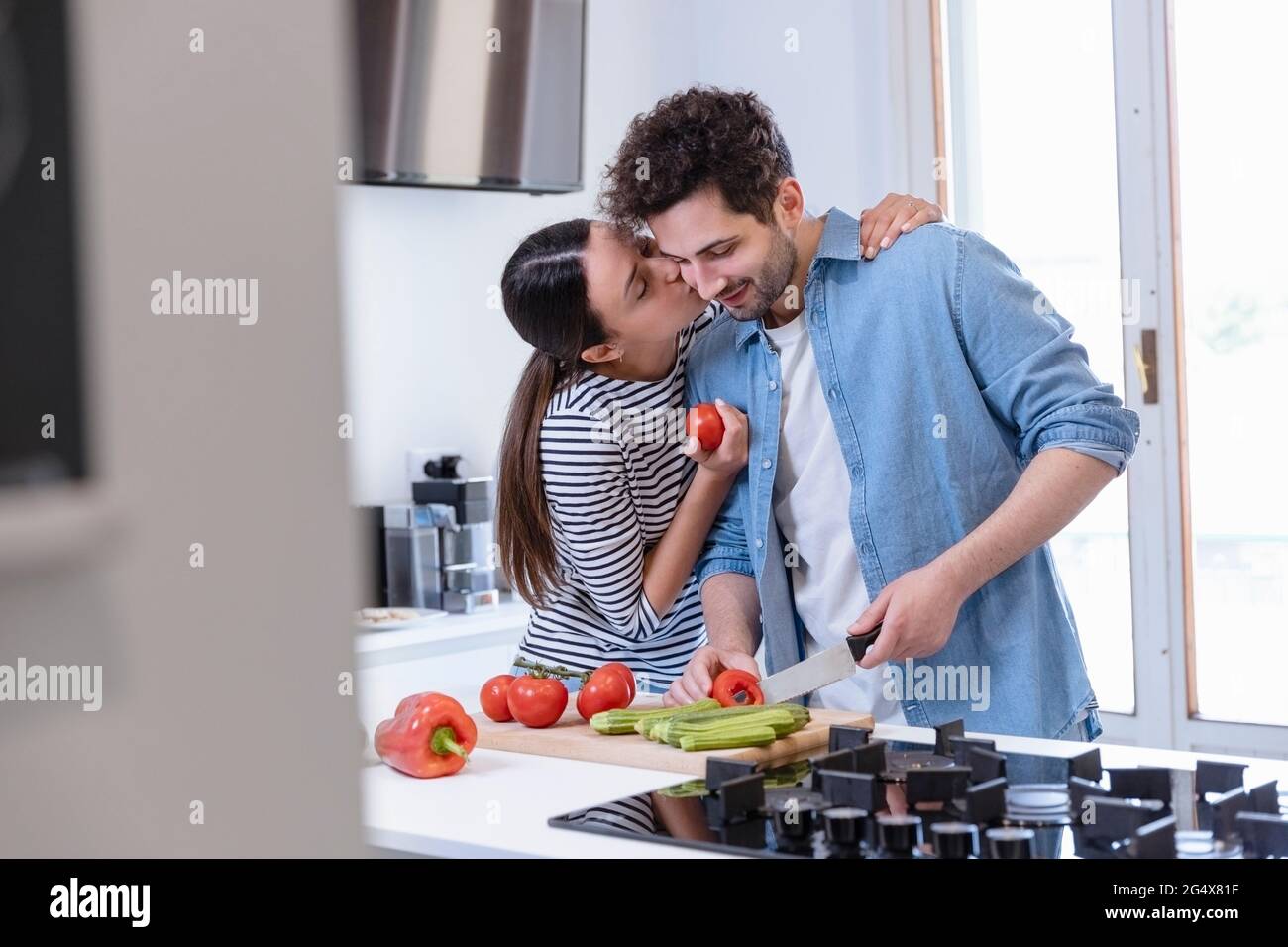 Young woman kissing boyfriend on cheek while cutting bell pepper in domestic kitchen Stock Photo