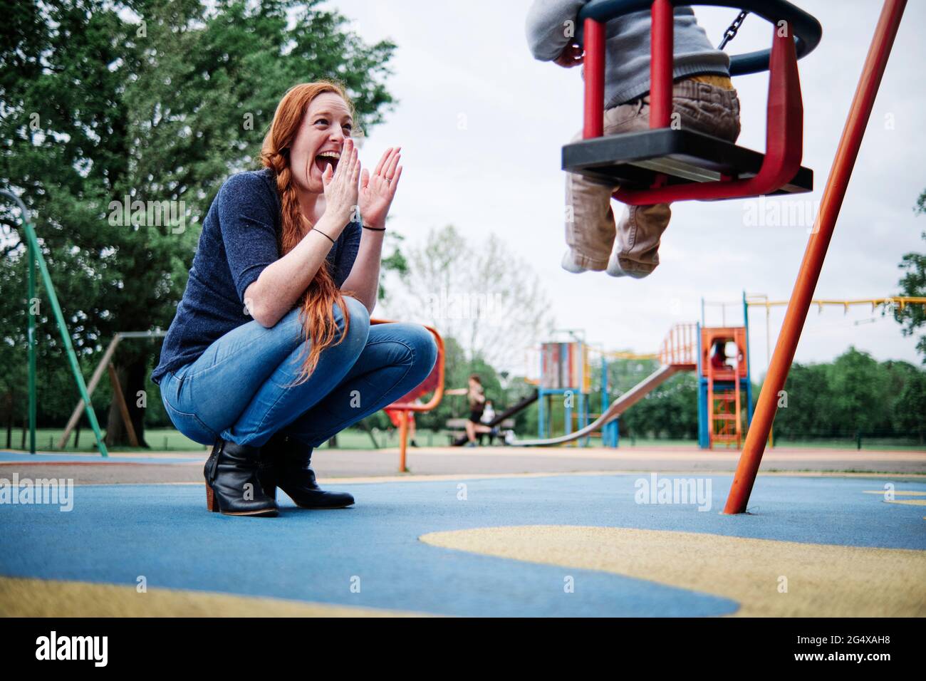 Happy mother clapping while crouching near son sitting on swing in playground Stock Photo