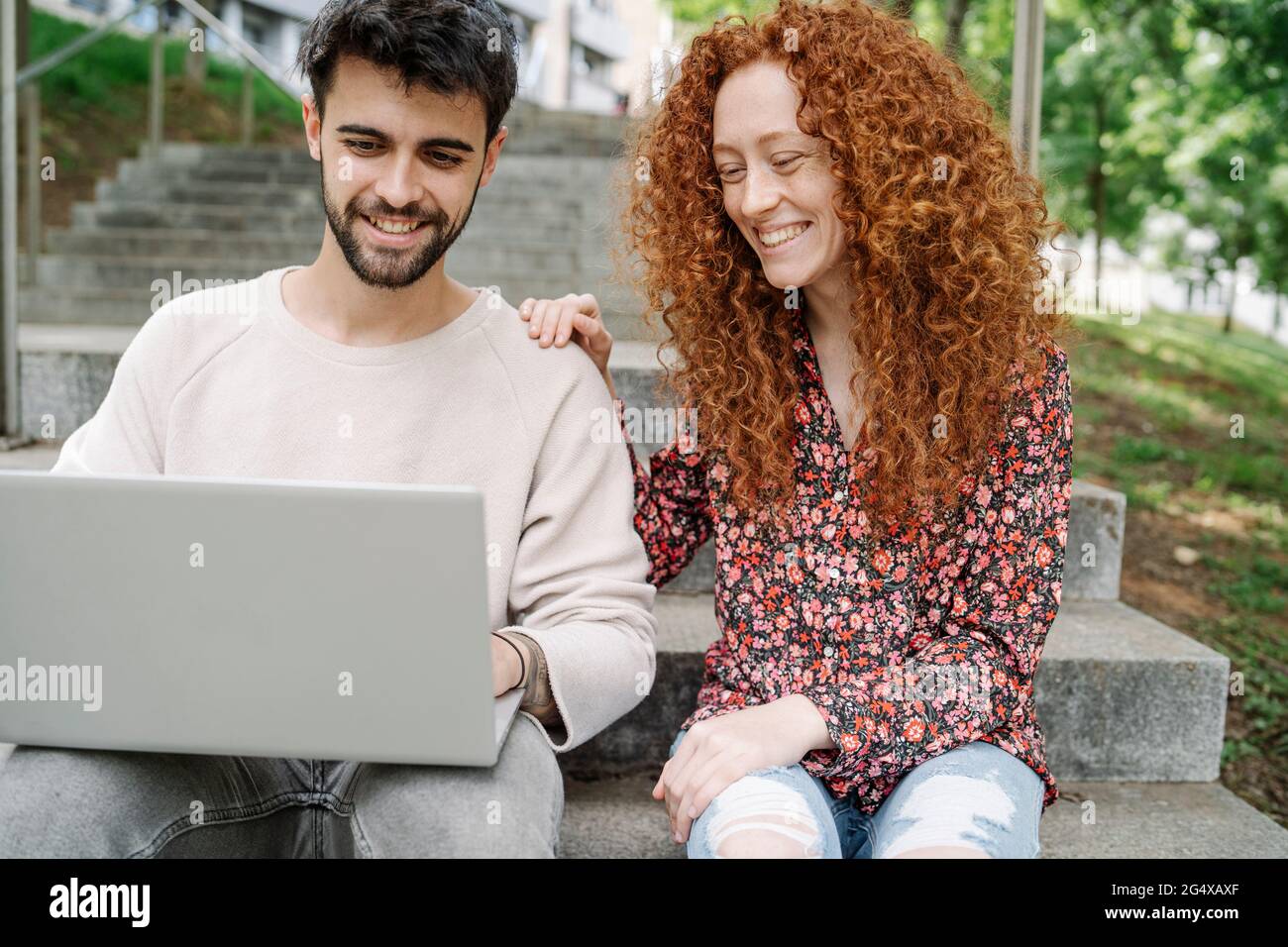 Smiling young couple using laptop while sitting on steps in public park Stock Photo