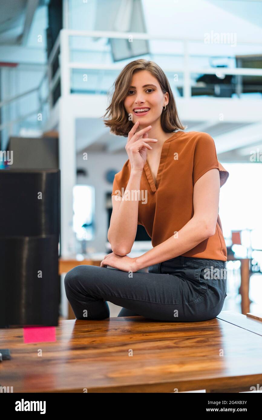 Smiling young professional sitting on desk in creative office Stock Photo
