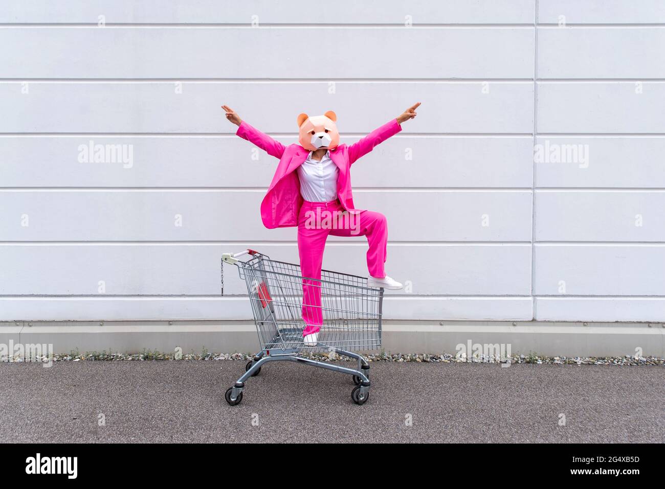 Woman wearing vibrant pink suit and bear mask standing in shopping cart with raised arms Stock Photo