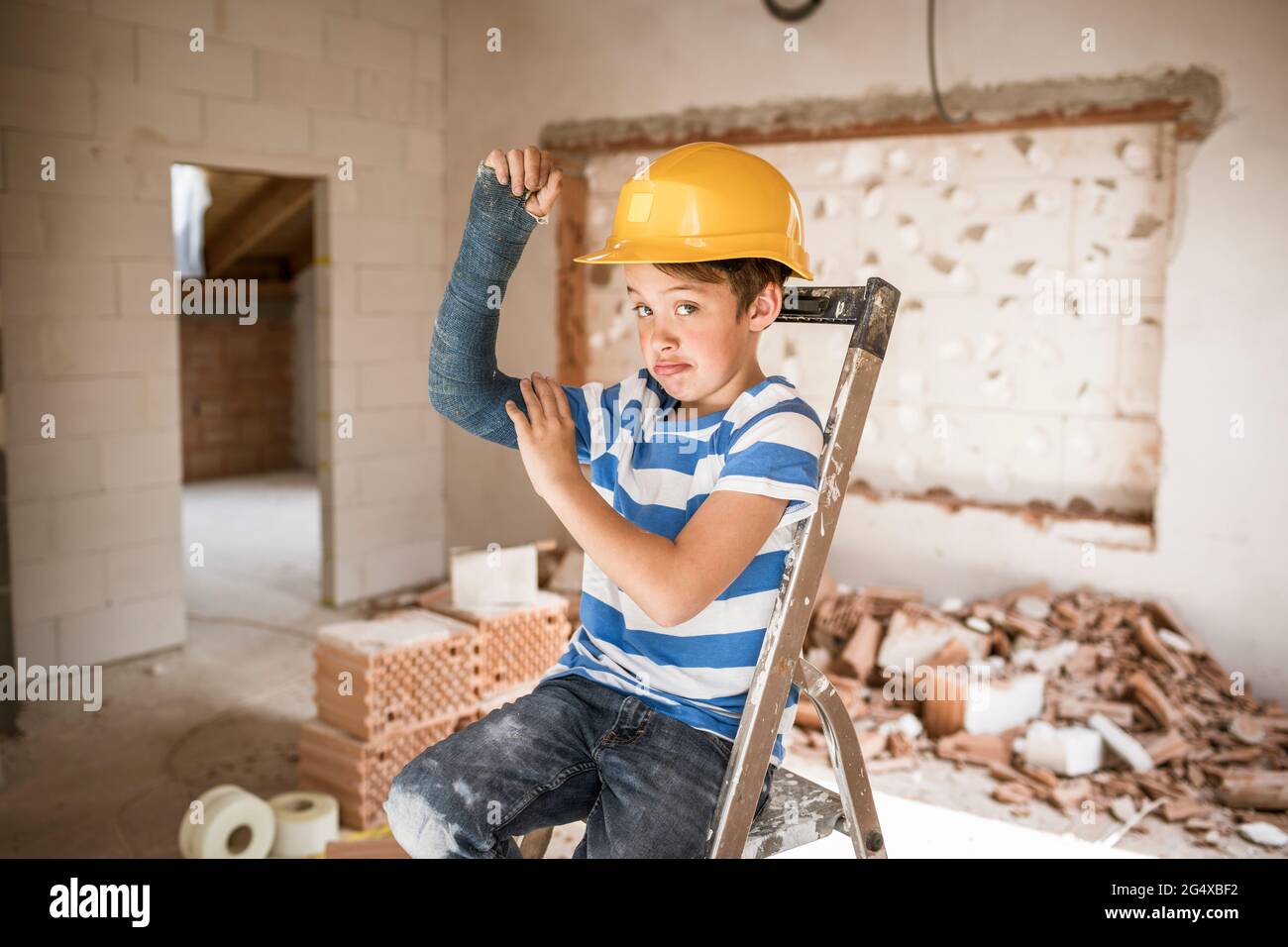 Boy flexing broken arm muscles while sitting on ladder during house renovation Stock Photo