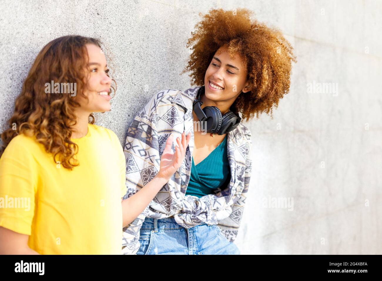 Young female friends talking while standing in front of wall Stock Photo