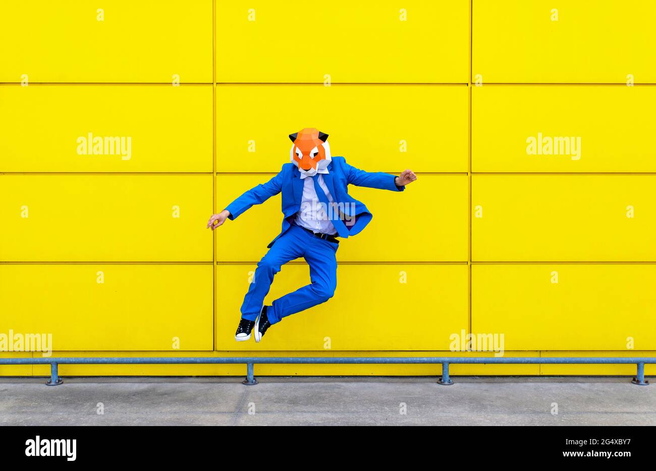 Man wearing vibrant blue suit and tiger mask jumping against yellow wall Stock Photo