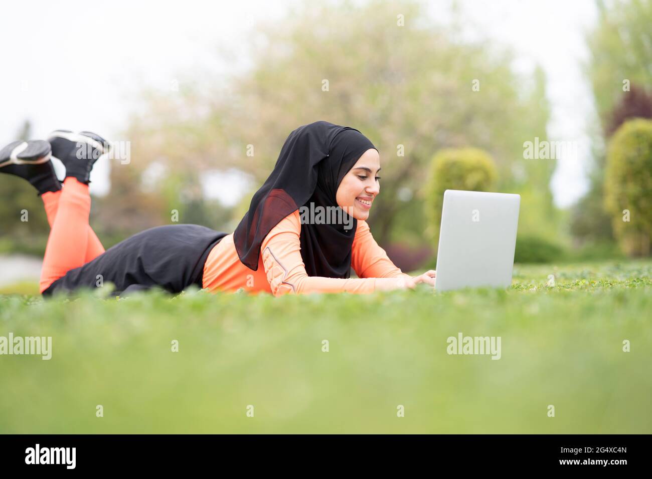 Young Arab woman lying on grass while using laptop in park Stock Photo