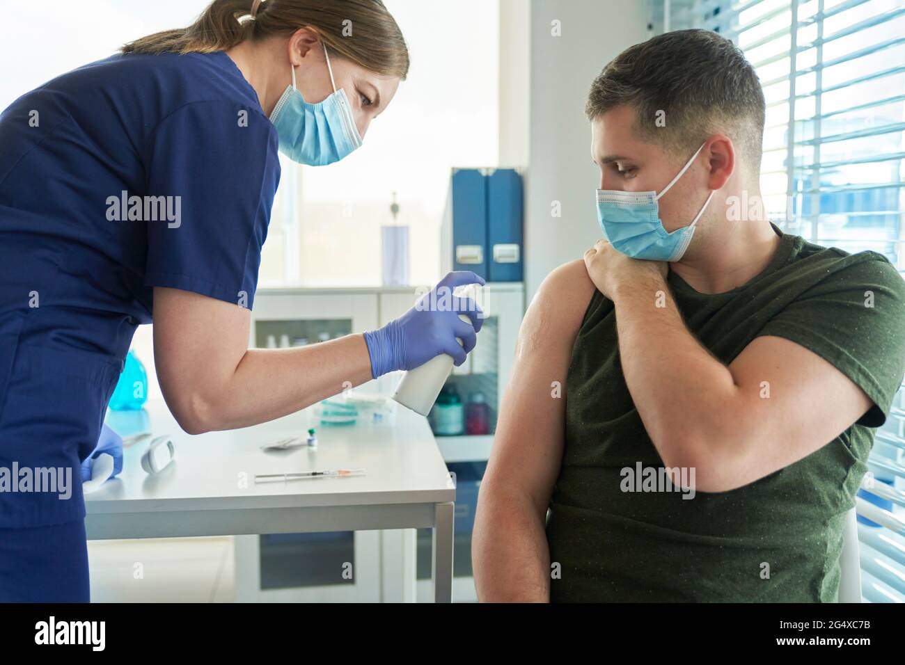 Female doctor spraying disinfectant on arm of male patient before giving COVID-19 vaccine at clinic Stock Photo